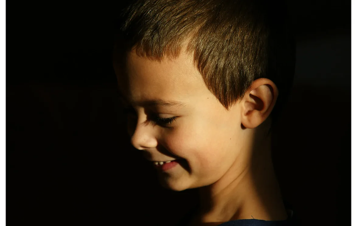 A child photographed in profile, lit only by soft daylight against a dark background